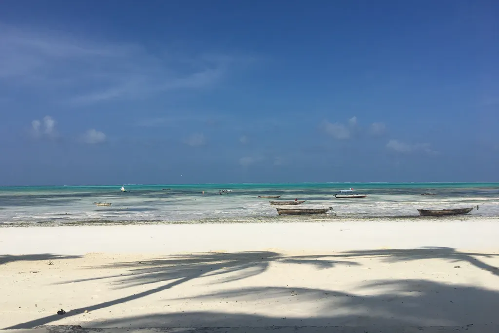Strand von Jambiani bei Ebbe mit Palmenschatten im Sand und Booten im seichten Wasser