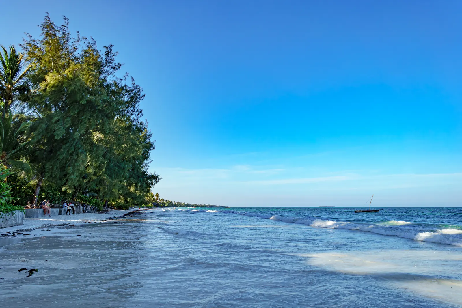 Sandstrand bei Nungwi mit Bäumen am Ufer, Wellen und einem Segelboot auf dem Meer